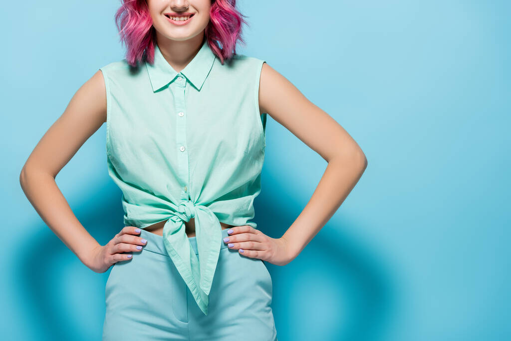 oříznutý pohled na mladou ženu s růžovými vlasy a rukama na bocích na modrém pozadí - Fotografie, Obrázek