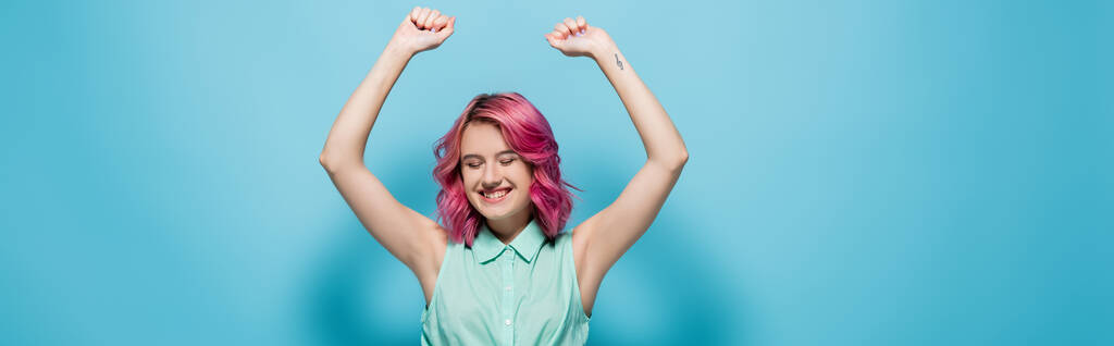 junge Frau mit rosafarbenen Haaren und lächelnden Händen in der Luft auf blauem Hintergrund, Panoramaaufnahme - Foto, Bild