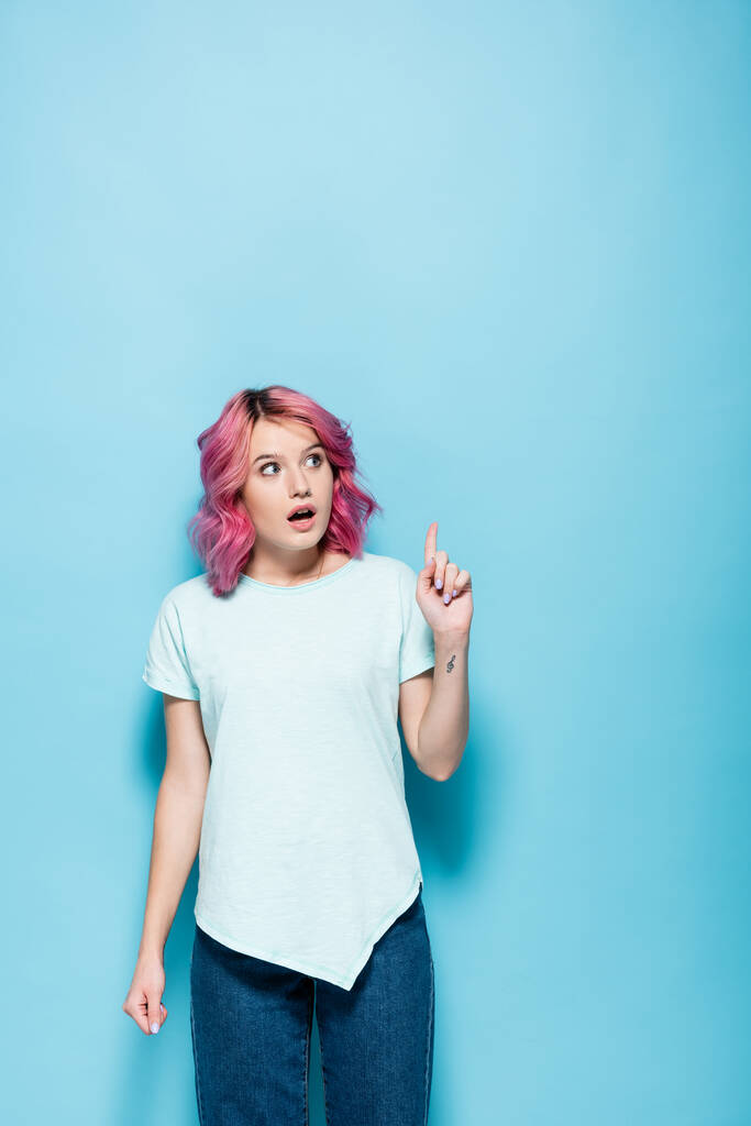zaskoczona młoda kobieta z różowymi włosami skierowana do góry na niebieskim tle - Zdjęcie, obraz