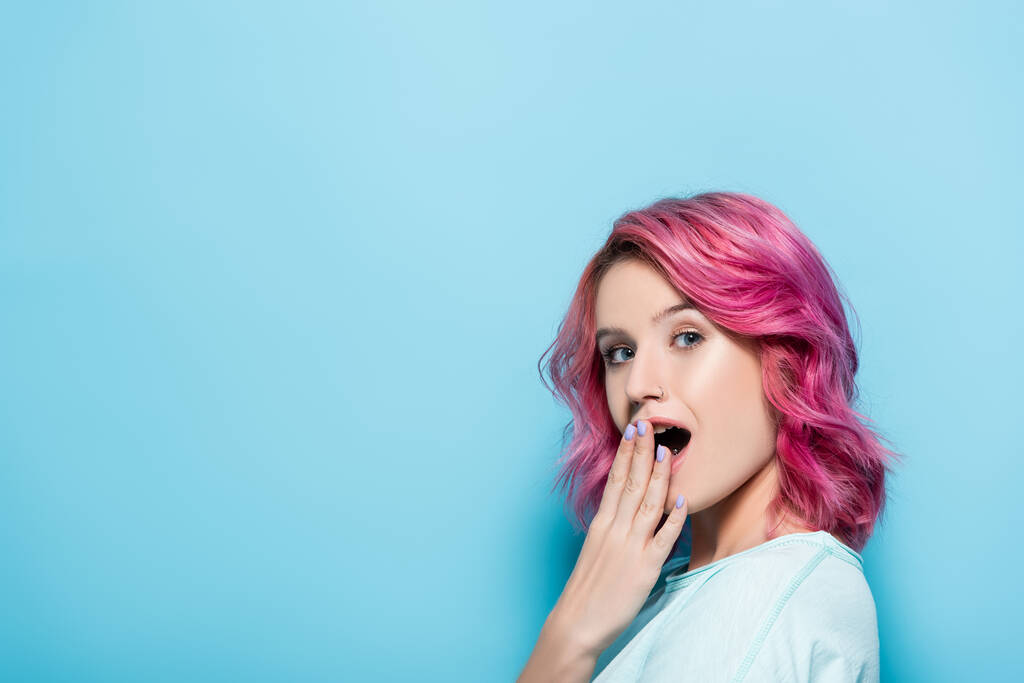 шокированная молодая женщина с розовыми волосами, покрывающими рот на синем фоне - Фото, изображение