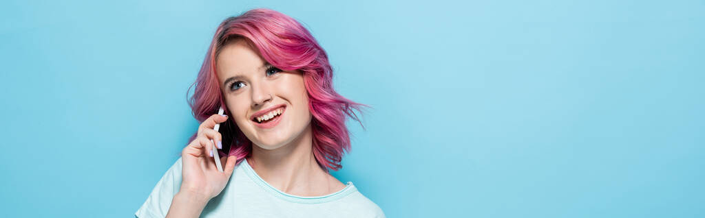 mujer joven con el pelo rosa hablando en el teléfono inteligente y sonriendo sobre fondo azul, tiro panorámico - Foto, Imagen