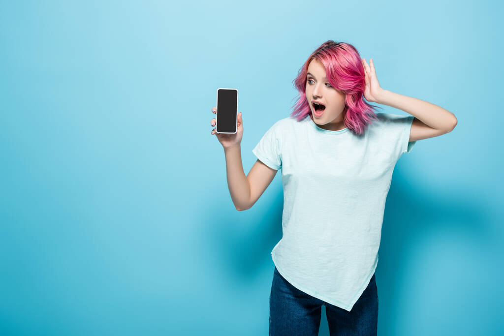 schockierte junge Frau mit rosa Haaren hält Smartphone mit leerem Bildschirm auf blauem Hintergrund - Foto, Bild