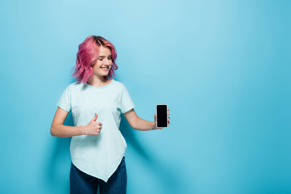 młoda kobieta z różowymi włosami trzyma smartfon z pustym ekranem i pokazuje kciuk w górę na niebieskim tle - Zdjęcie, obraz