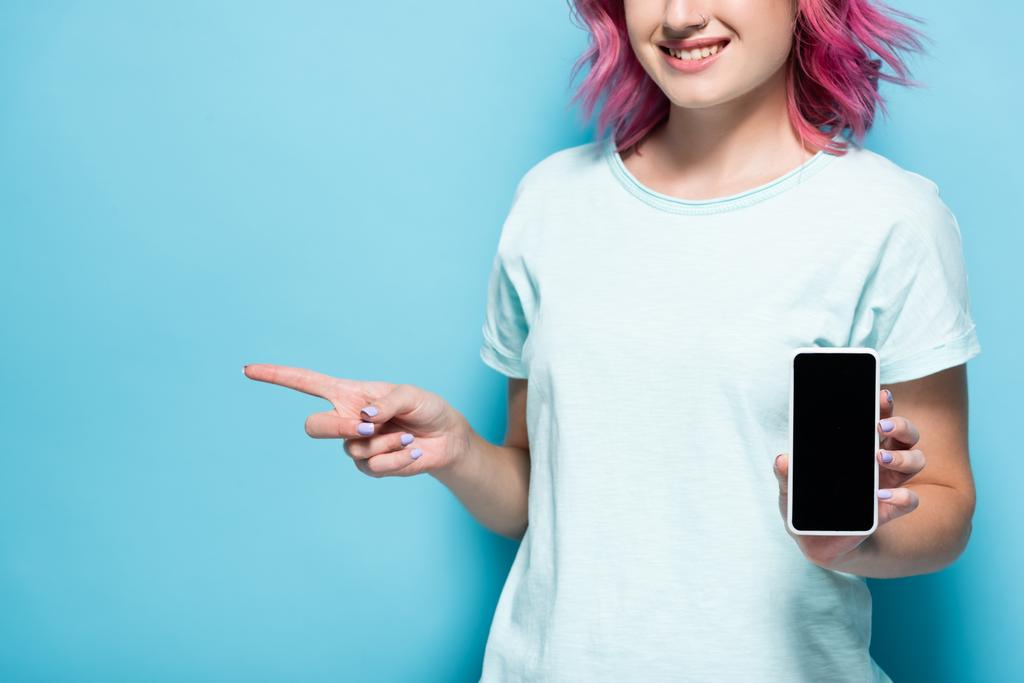 vista recortada de mujer joven con el pelo rosa sosteniendo teléfono inteligente con pantalla en blanco y apuntando a un lado sobre fondo azul - Foto, imagen