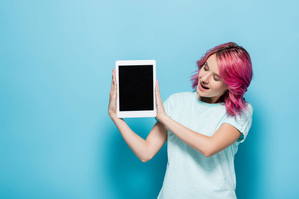 überraschte junge Frau mit rosa Haaren präsentiert digitales Tablet mit leerem Bildschirm auf blauem Hintergrund - Foto, Bild