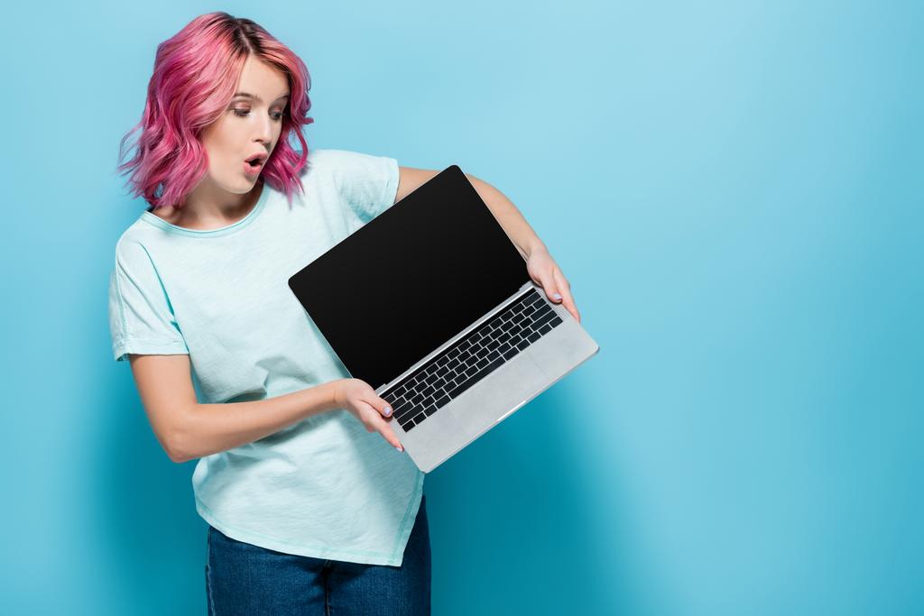 šokovaná mladá žena s růžovými vlasy ukazující notebook s prázdnou obrazovkou na modrém pozadí - Fotografie, Obrázek