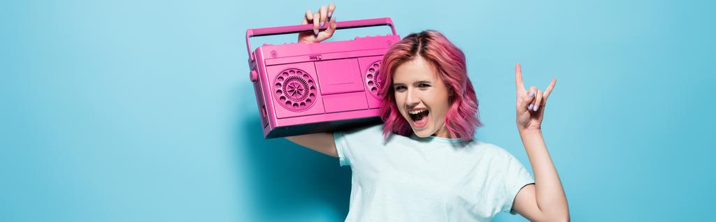 excited young woman with pink hair holding vintage tape recorder and showing rock sign on blue background, panoramic shot - Photo, Image