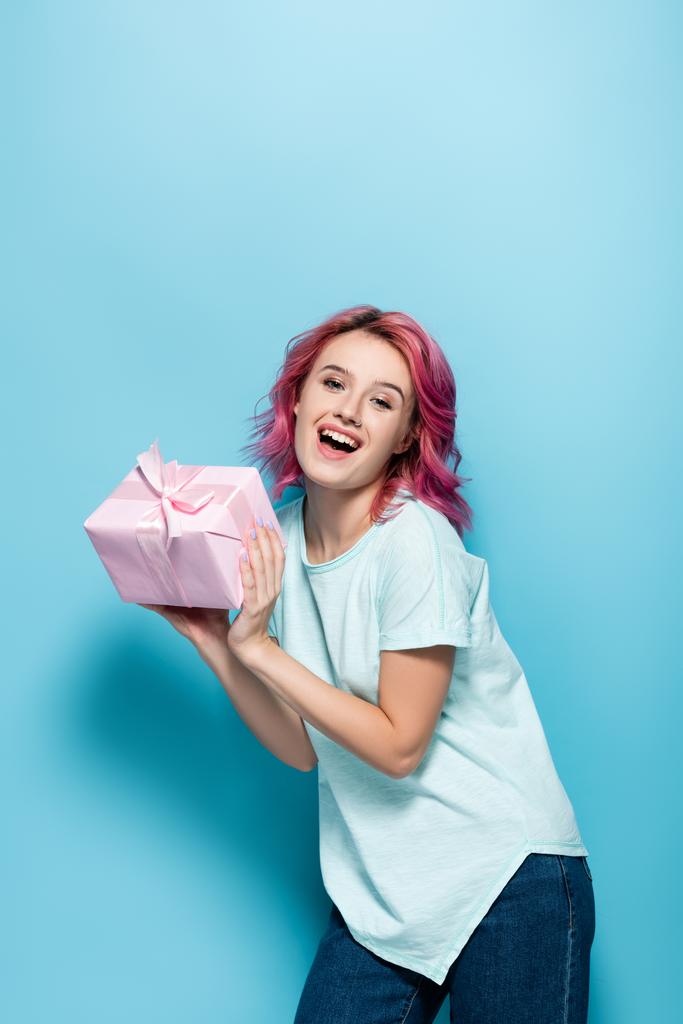 aufgeregte junge Frau mit rosa Haaren hält Geschenkbox mit Schleife auf blauem Hintergrund - Foto, Bild