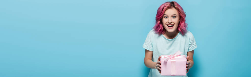 excité jeune femme avec des cheveux roses tenant boîte cadeau avec arc sur fond bleu, plan panoramique - Photo, image