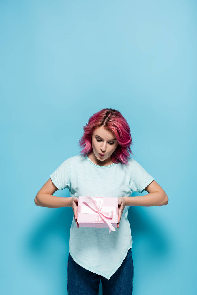 mujer joven sorprendida con cabello rosa sosteniendo caja de regalo con lazo sobre fondo azul - Foto, Imagen