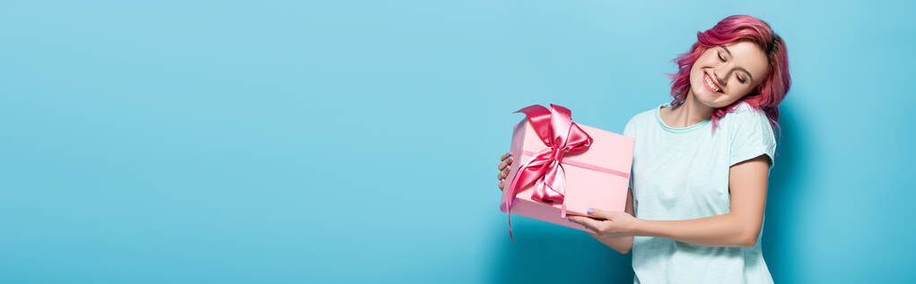 молодая женщина с розовыми волосами держа подарочную коробку с луком и улыбаясь с закрытыми глазами на голубом фоне, панорамный снимок - Фото, изображение