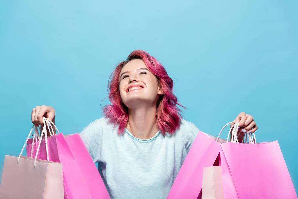 młoda kobieta z różowymi włosami trzyma torby na zakupy i uśmiecha się odizolowany na niebiesko - Zdjęcie, obraz