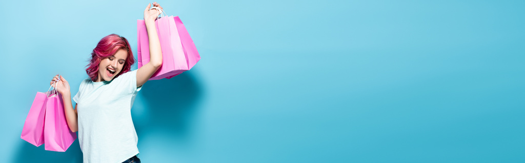 jovem com cabelo rosa segurando sacos de compras e sorrindo no fundo azul, tiro panorâmico - Foto, Imagem