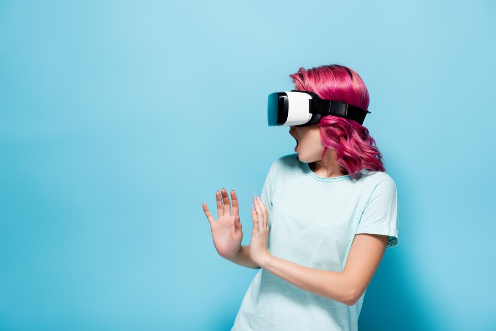 verängstigte junge Frau mit rosa Haaren im vr-Headset gestikulierend auf blauem Hintergrund - Foto, Bild