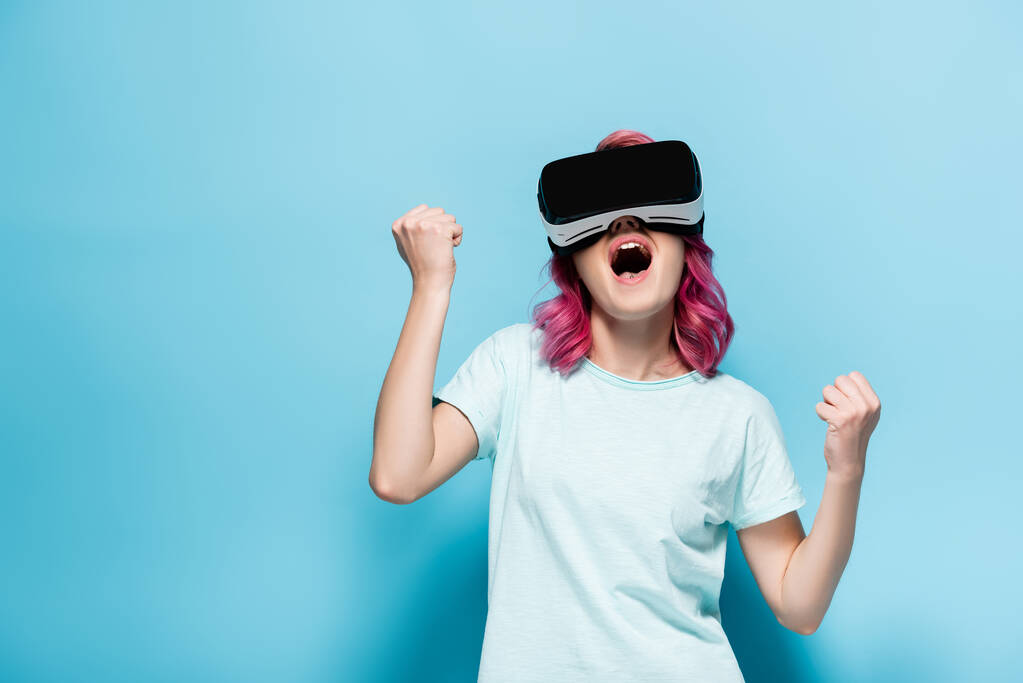 возбужденная молодая женщина с розовыми волосами в VR гарнитуре показывая да жест на голубом фоне - Фото, изображение