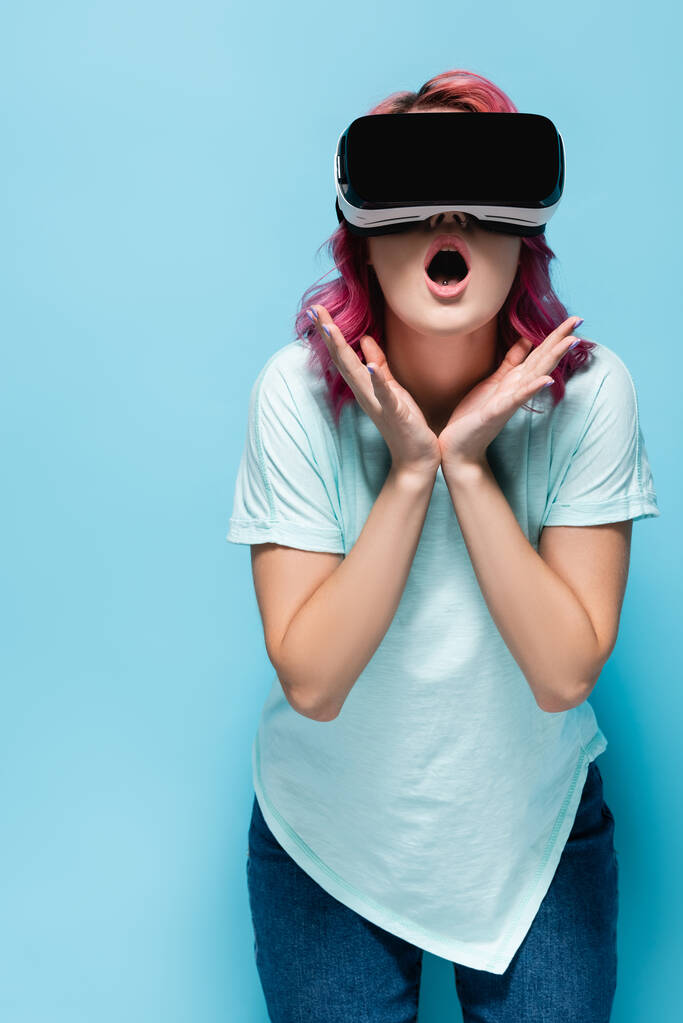 chocado jovem mulher com cabelo rosa no vr headset segurando as mãos perto do rosto no fundo azul - Foto, Imagem