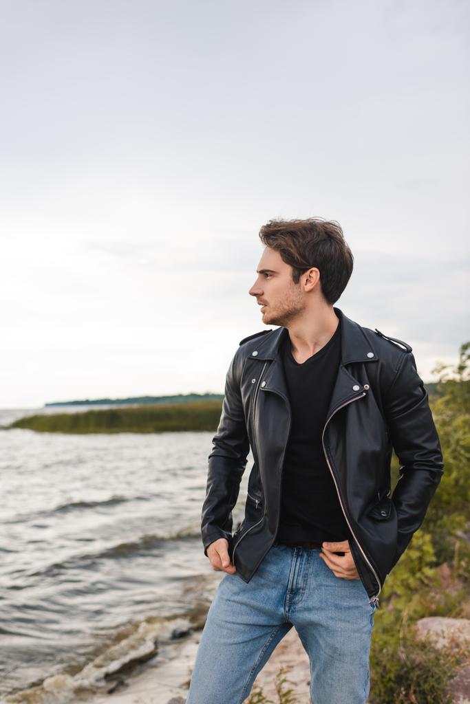 Young man in jeans and leather jacket looking away on sea coast  - Photo, Image
