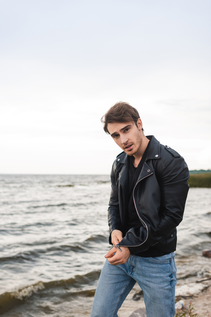 Mladý muž v džínách a kožené bundě při pohledu na kameru na pláži  - Fotografie, Obrázek