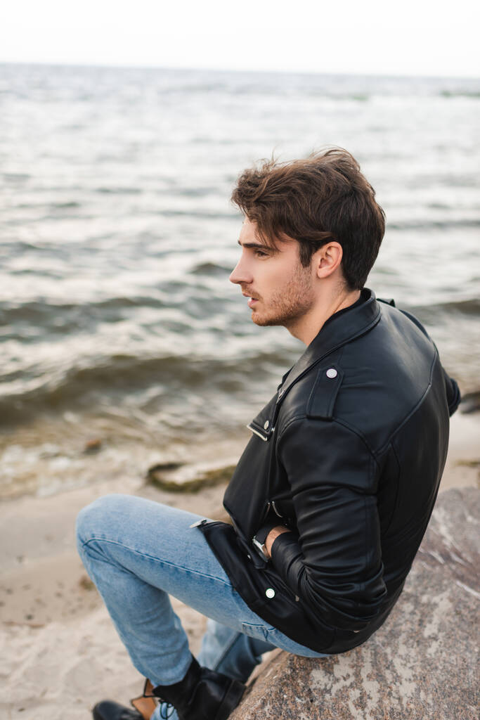 Mann in Lederjacke und Jeans sitzt auf Stein am Strand  - Foto, Bild