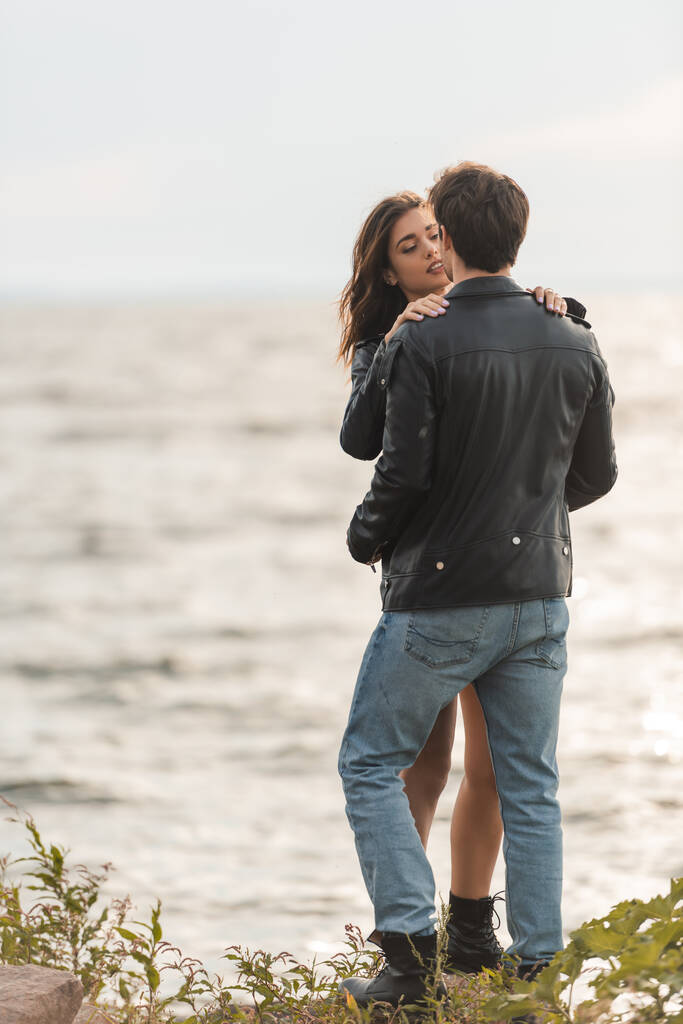 Mulher morena abraçando namorado em jaqueta de couro e jeans na praia perto do mar  - Foto, Imagem