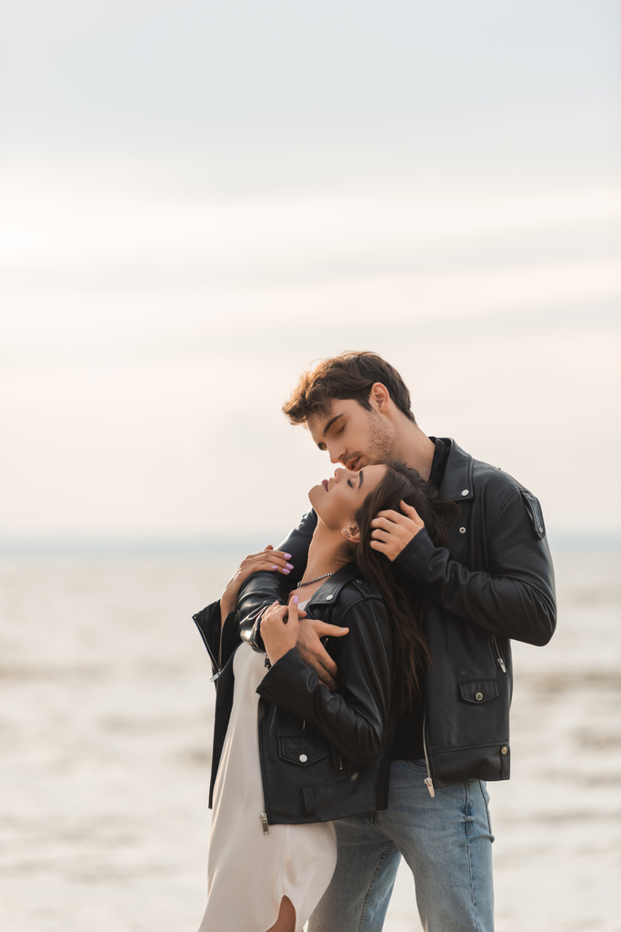 Mladý muž objímání a dotýkání vlasů přítelkyně v šatech a kožené bundy v blízkosti moře - Fotografie, Obrázek