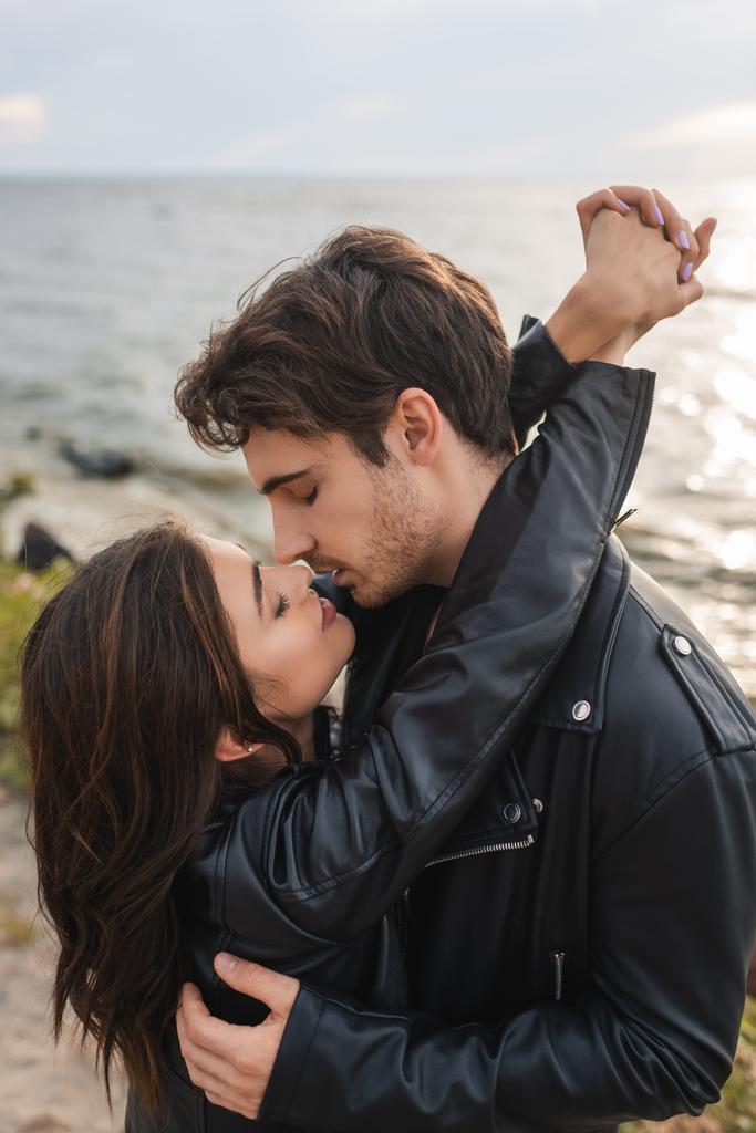Mann küsst und umarmt brünette Freundin in Lederjacke am Strand  - Foto, Bild