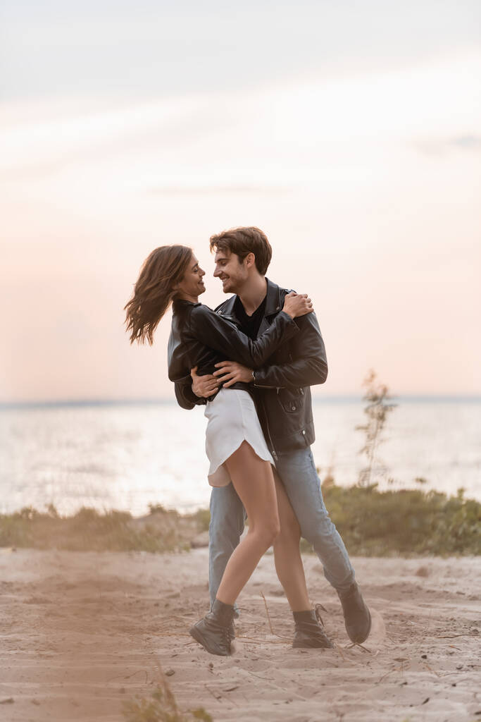 Selective focus of man in leather jacket embracing girlfriend on beach at sunset  - Photo, Image