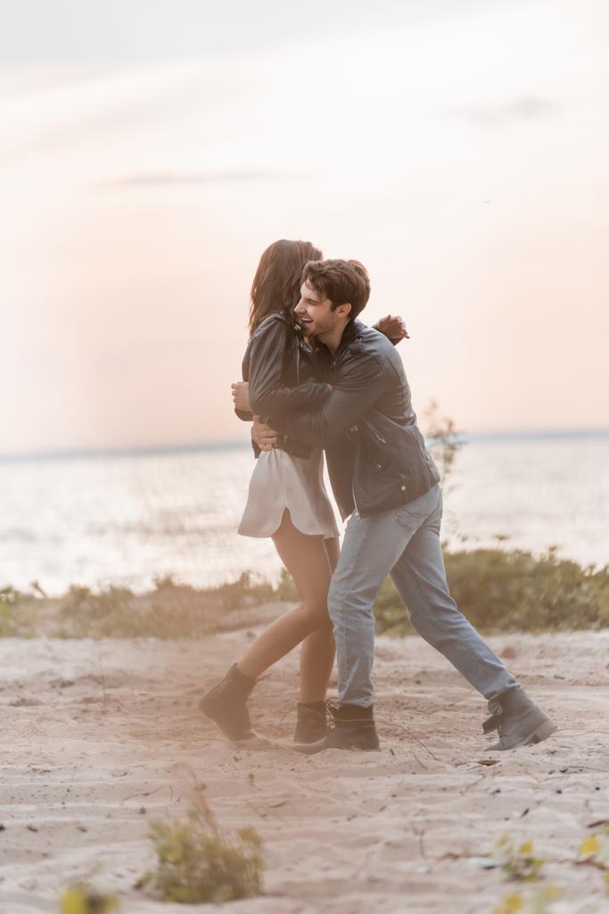 Focus selettivo dell'uomo in giacca di pelle che abbraccia la ragazza in abito sulla spiaggia al tramonto  - Foto, immagini