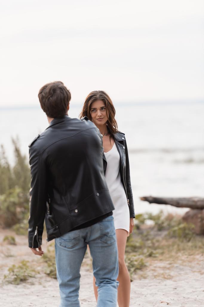 Selective focus of young woman in dress and leather jacket looking at boyfriend on beach  - Photo, Image