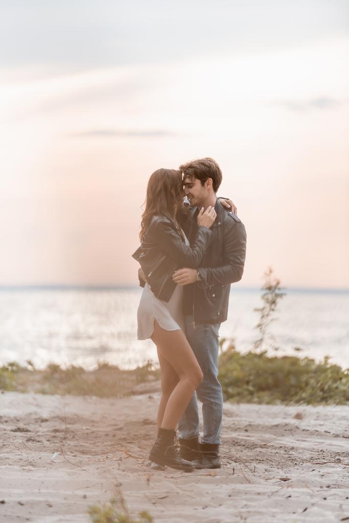 Selektywne skupienie młodego mężczyzny obejmującego dziewczynę w sukience i skórzanej kurtce na plaży o zachodzie słońca  - Zdjęcie, obraz