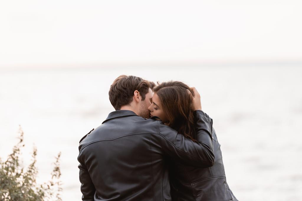 Rückansicht eines jungen Paares in Lederjacken, das sich am Meer küsst  - Foto, Bild
