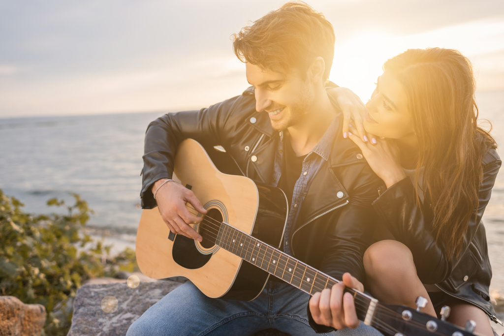 Młody mężczyzna w skórzanej kurtce gra na gitarze akustycznej w pobliżu dziewczyny na plaży o zachodzie słońca  - Zdjęcie, obraz