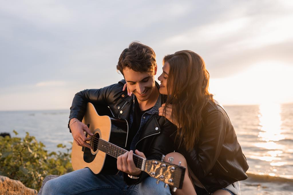 Selective focus of woman in leather jacket embracing boyfriend playing acoustic guitar during sunset on beach  - Photo, Image