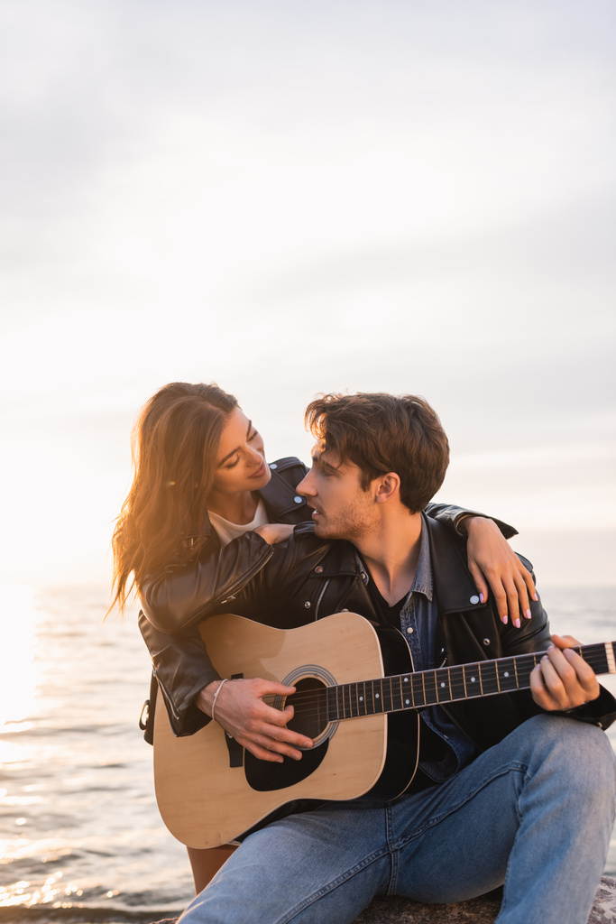 Junge Frau in Lederjacke umarmt Freund, der bei Sonnenuntergang am Meer Akustikgitarre spielt  - Foto, Bild