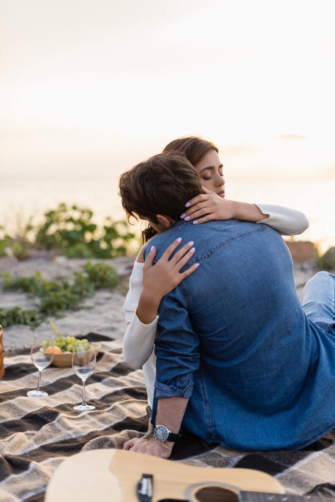 Selective focus of woman embracing boyfriend near acoustic guitar and wine glasses on beach  - Photo, Image