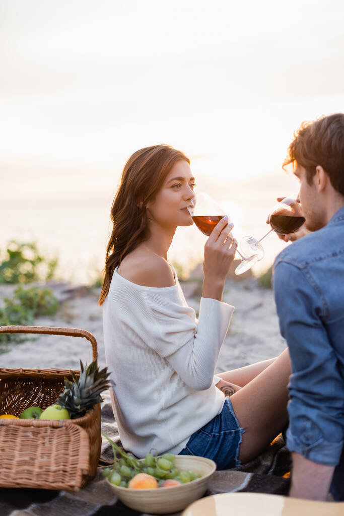 Foco seletivo da mulher bebendo vinho perto do namorado durante piquenique na praia ao pôr do sol  - Foto, Imagem