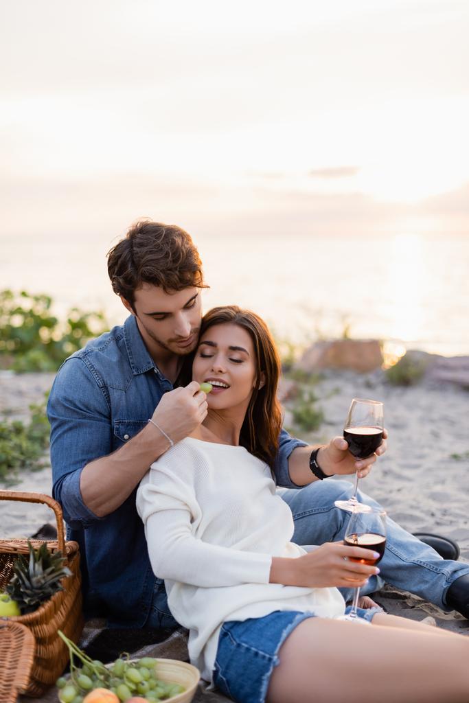 Enfoque selectivo del hombre alimentando a su novia con uva mientras sostiene una copa de vino en la playa  - Foto, Imagen