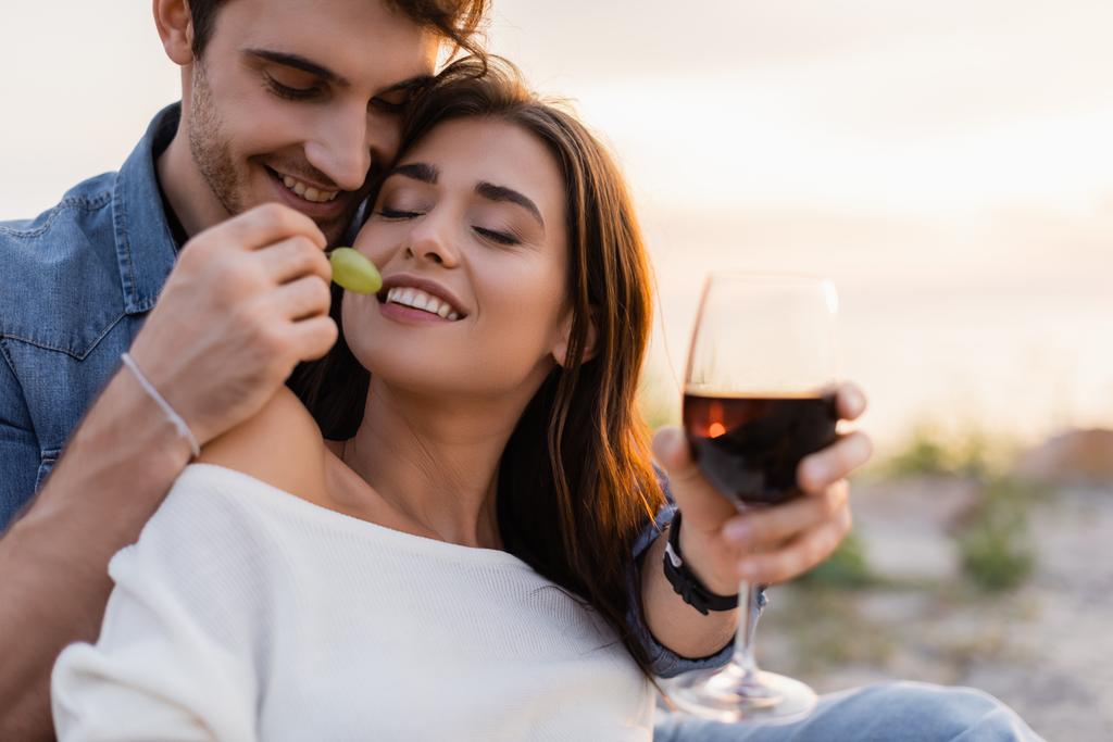 Enfoque selectivo del hombre alimentando a su novia con uva mientras sostiene una copa de vino en la playa  - Foto, imagen