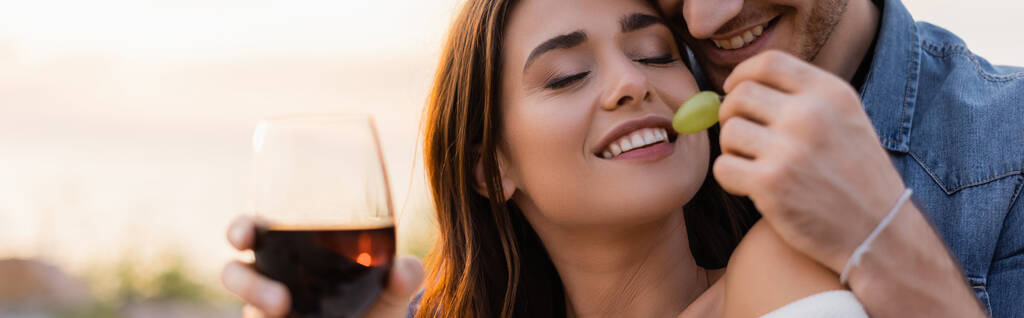 Вебсайт заголовок человека с бокалом вина кормящей подруги с виноградом на пляже вечером  - Фото, изображение