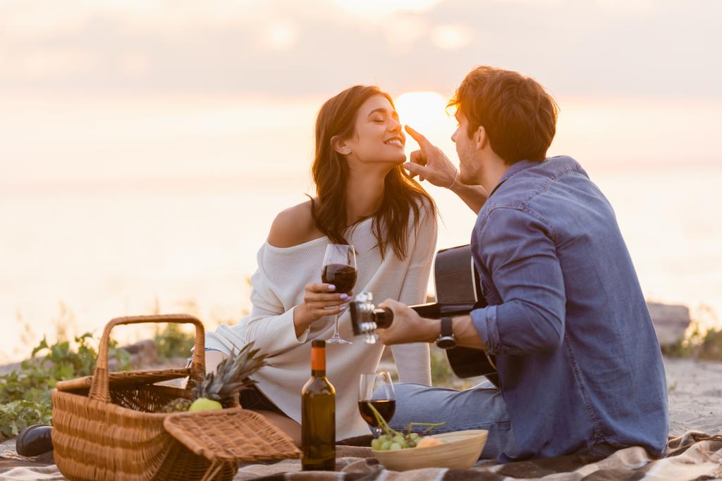 男とともにアコースティックギタータッチ鼻のガールフレンドとともにガラスのワインでビーチで日没  - 写真・画像