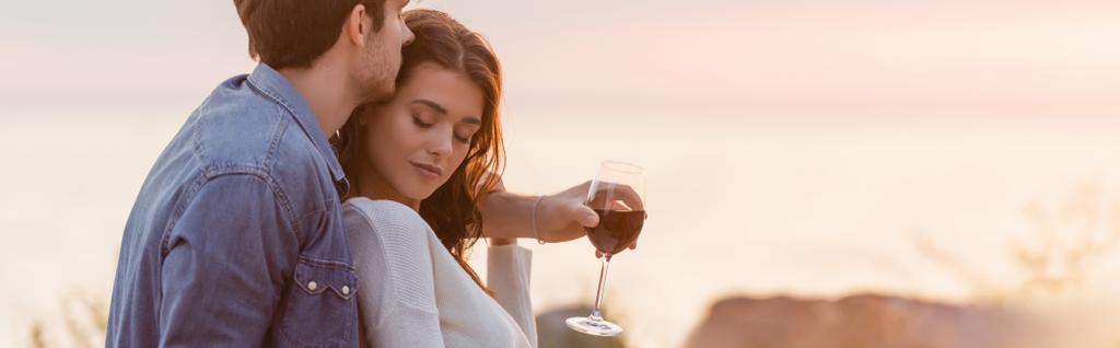 Honlap fejléce férfi ölelés nő, miközben kezében pohár bor a strandon naplementekor  - Fotó, kép