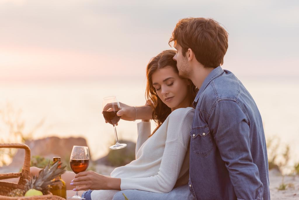 Enfoque selectivo del hombre abrazando a la novia con una copa de vino durante el picnic en la playa  - Foto, imagen