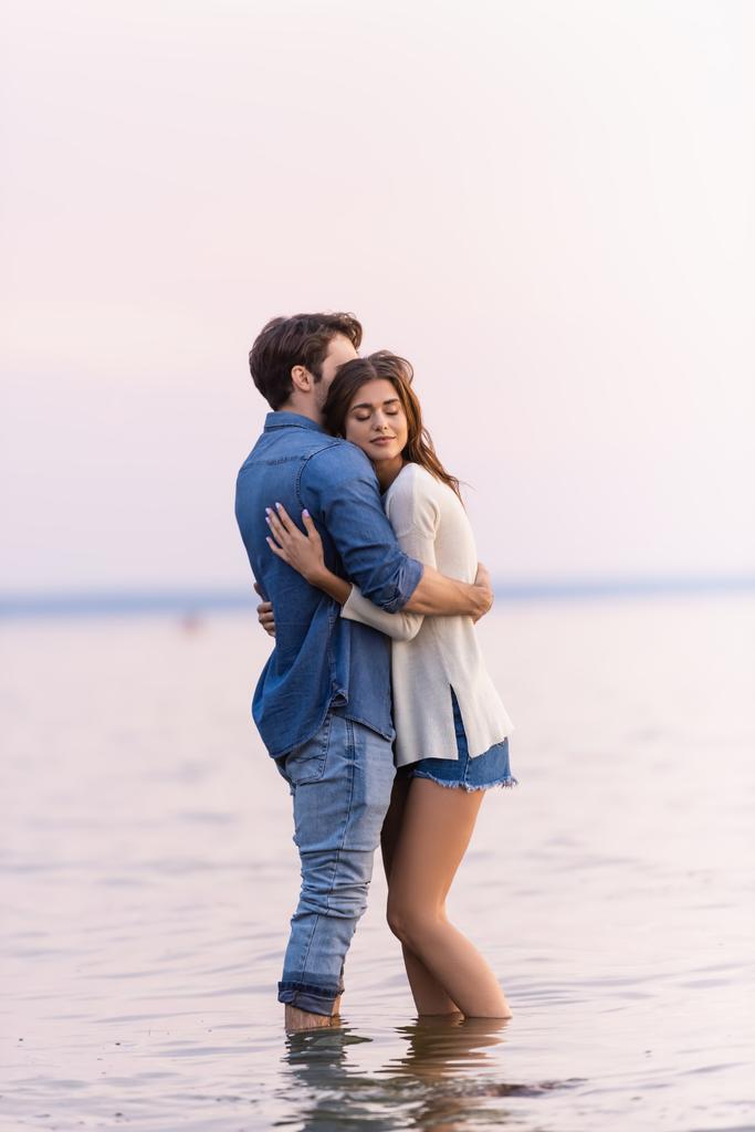 Side view of brunette woman with closed eyes hugging boyfriend while standing in sea  - Photo, Image