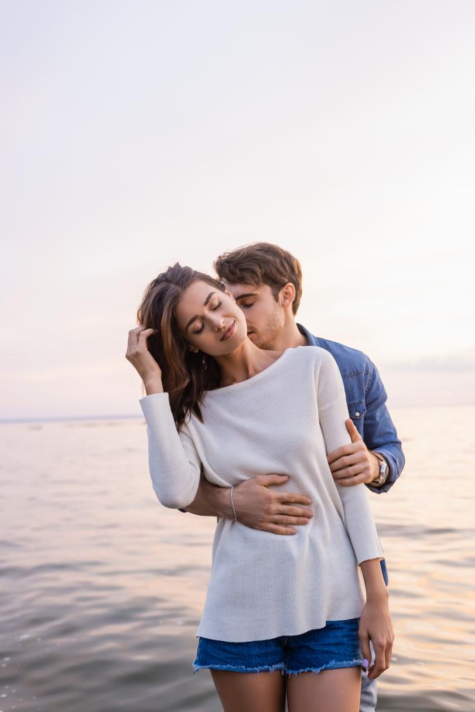 Junger Mann küsst Hals und umarmt brünette Frau am Meer  - Foto, Bild