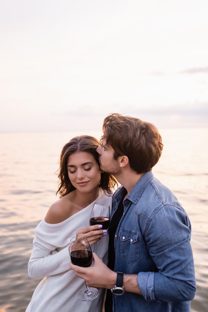 Νεαρός φιλάει τη φίλη του με ένα ποτήρι κρασί κοντά στη θάλασσα το βράδυ  - Φωτογραφία, εικόνα
