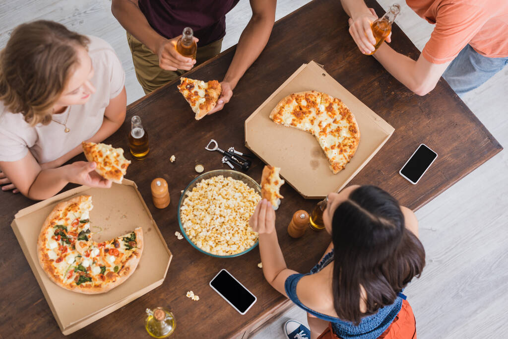 κορυφαία άποψη των πολυεθνικών φίλων πίνοντας μπύρα και τρώγοντας πίτσα κατά τη διάρκεια του πάρτι - Φωτογραφία, εικόνα