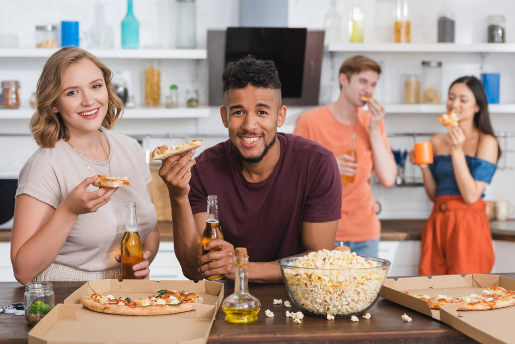zadowoleni wielonarodowi przyjaciele patrząc w kamerę podczas jedzenia pizzy podczas imprezy - Zdjęcie, obraz
