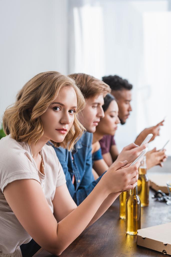 young woman looking at camera while chatting on smartphone near multicultural friends - Photo, Image