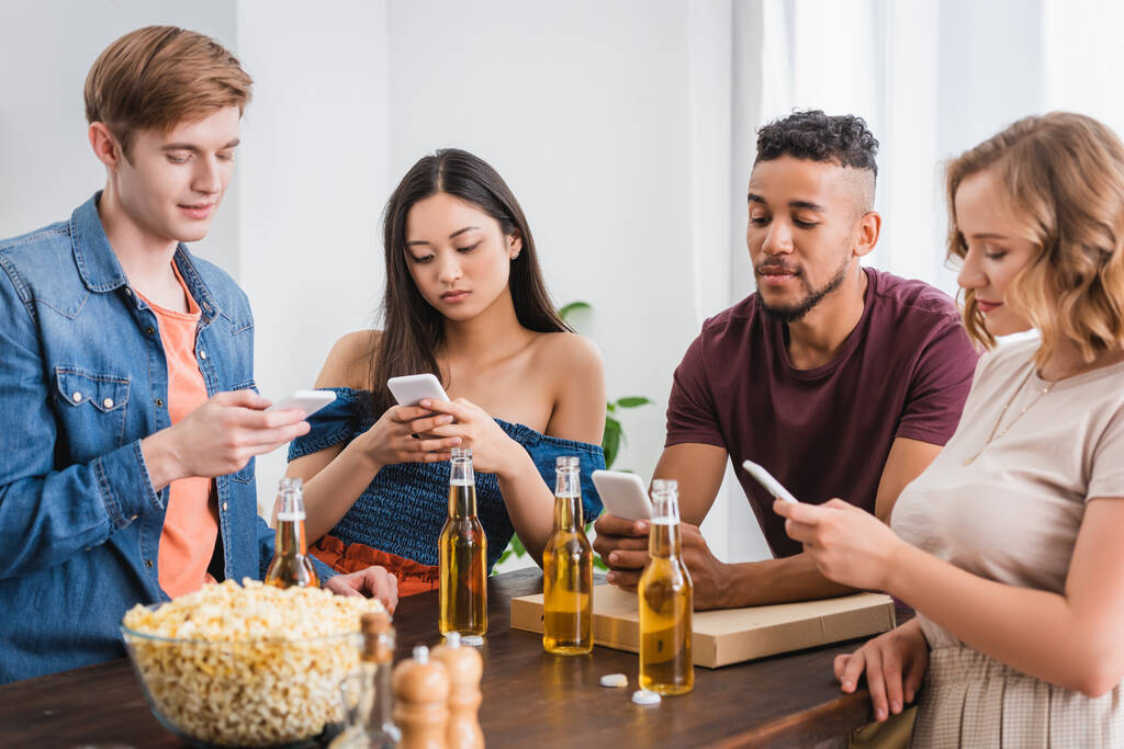 selectieve focus van multi-etnische vrienden berichten op smartphones in de buurt van bier en popcorn tijdens het feest - Foto, afbeelding