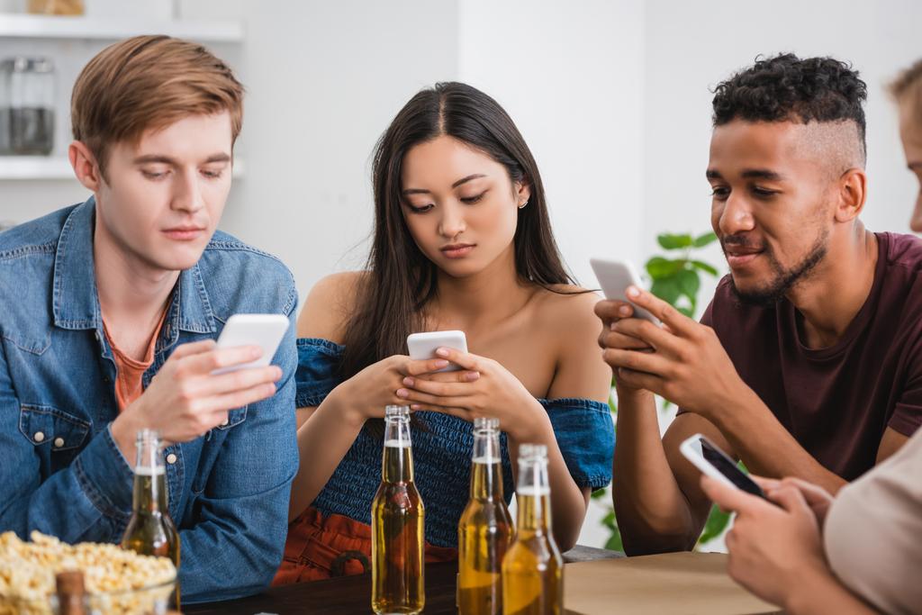enfoque selectivo de amigos multiculturales charlando en teléfonos inteligentes cerca de la cerveza durante la fiesta - Foto, Imagen